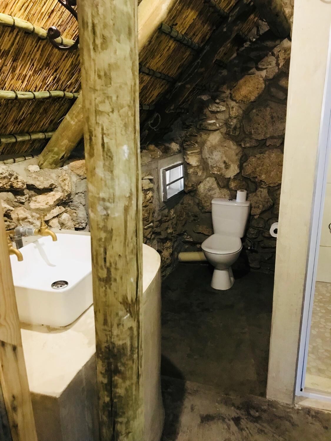 A rustic bathroom with a stone wall, a white toilet, and a wooden bathtub.
