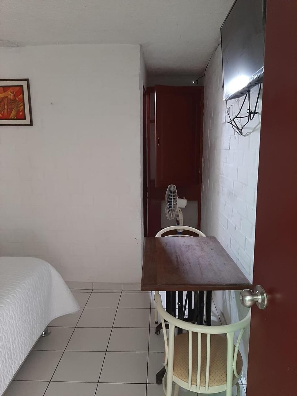 A small room with a bed, a table, and a chair. There is a wall-mounted fan and a picture frame on the wall.