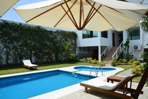A swimming pool with lounge chairs and an umbrella, surrounded by a garden and a modern building. - Qallwa Asia (Asia)