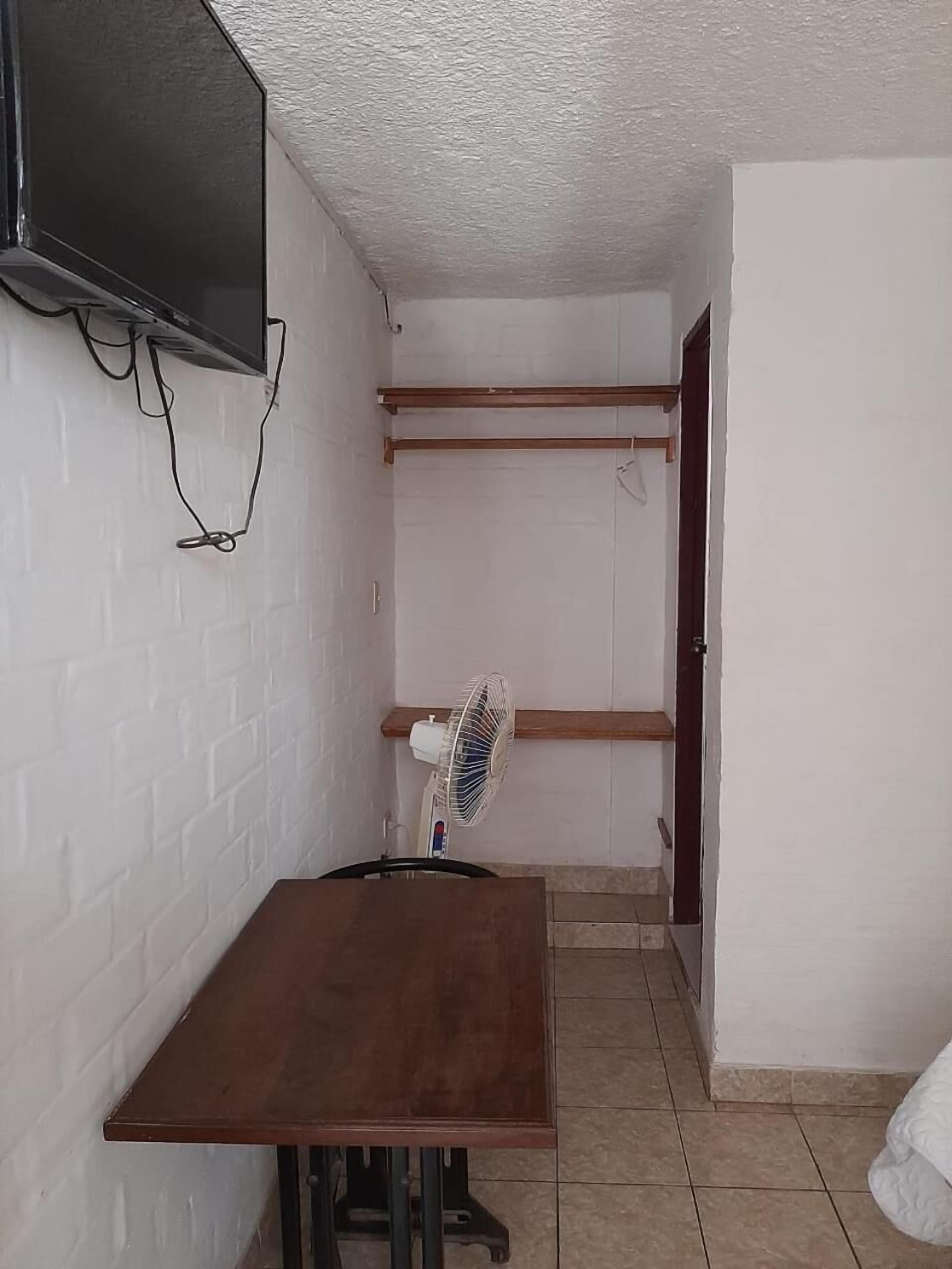 A small room with a wooden table, a white fan, a TV mounted on the wall, and a door.