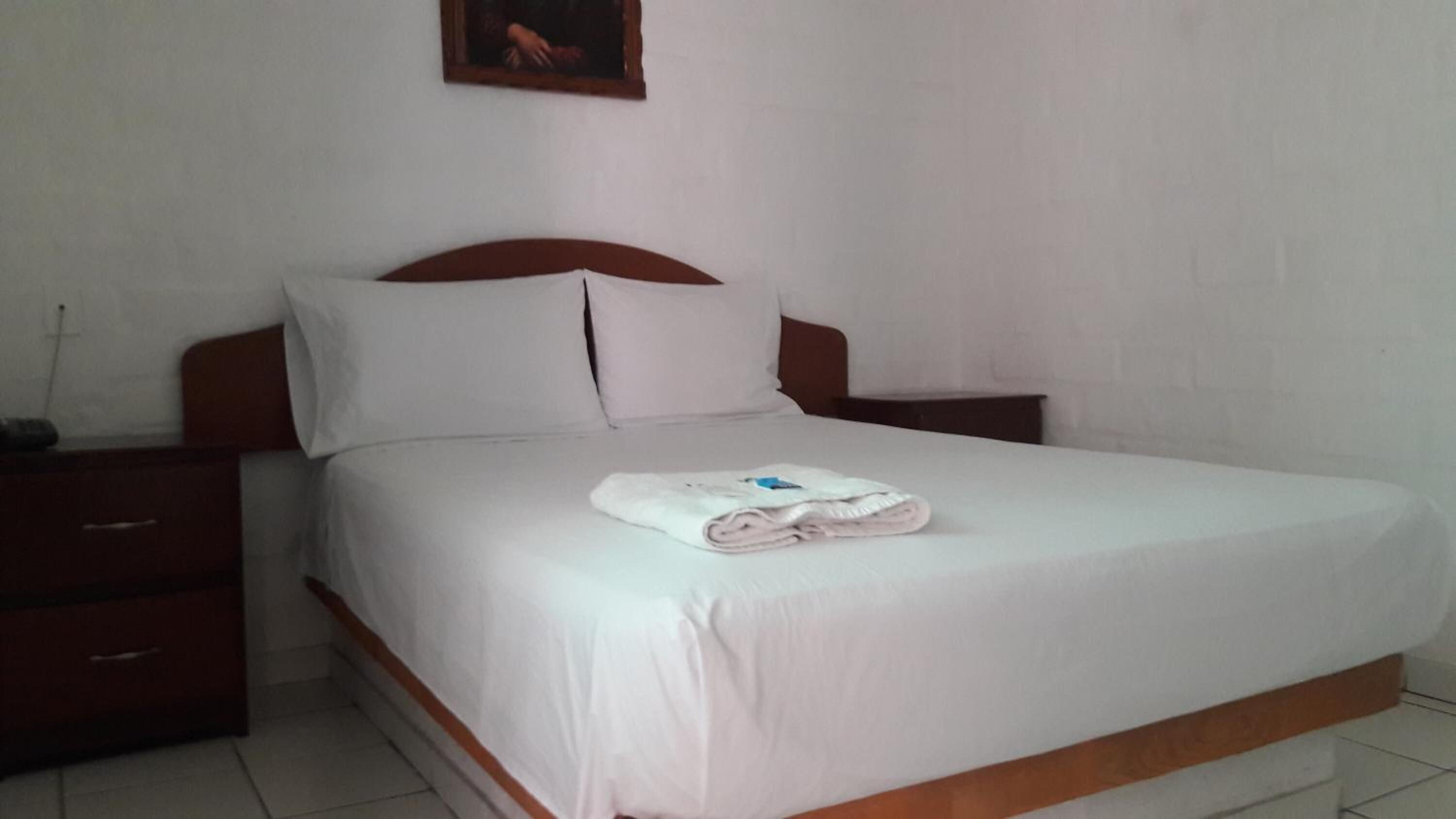 A neatly made bed with white linens and a folded towel on top, a wooden headboard, and a small bedside table with a lamp.