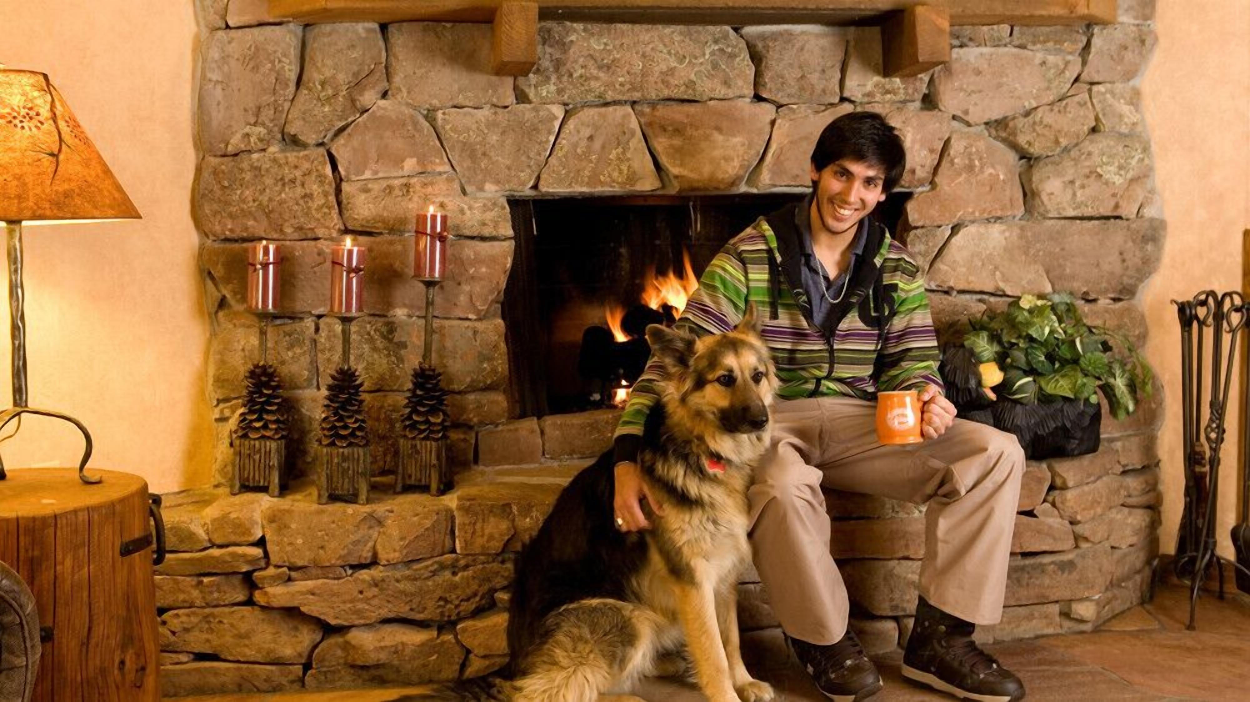 A person sitting next to a dog in front of a stone fireplace with a fire, surrounded by pine cones and candles.