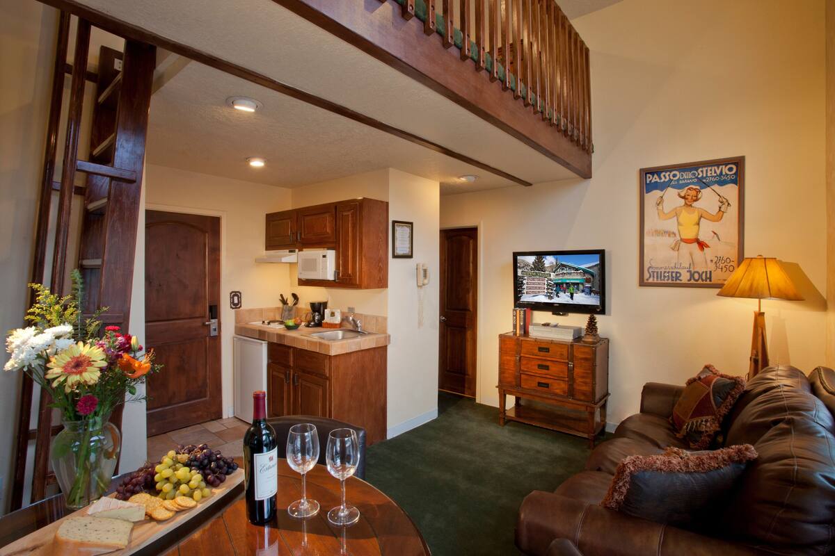A living room with a brown leather couch, a wooden coffee table with a bottle of wine and glasses, a TV on a wooden stand, and a flower vase.
