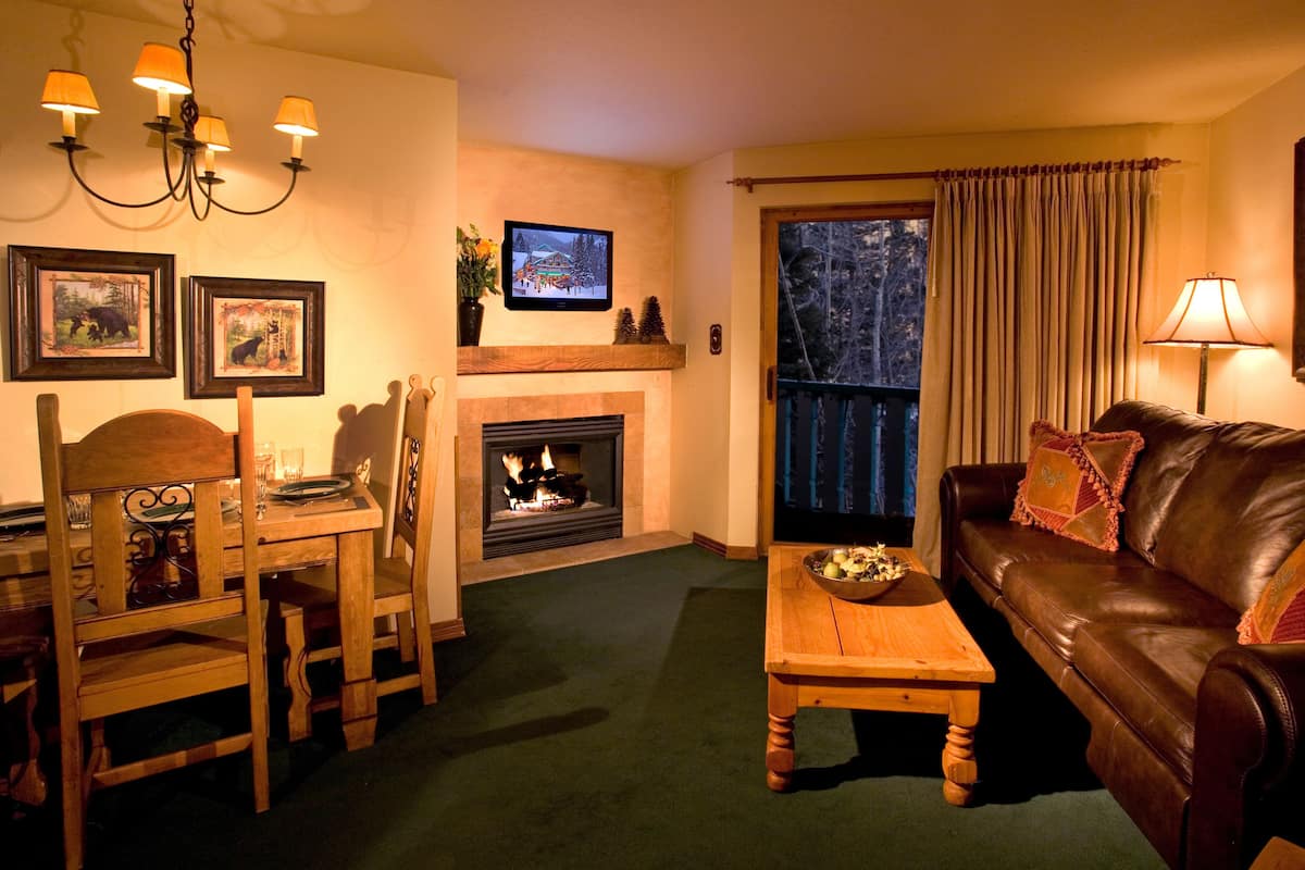 A cozy living room with a fireplace, a wooden dining table with chairs, a leather sofa, and a television mounted on the wall.
