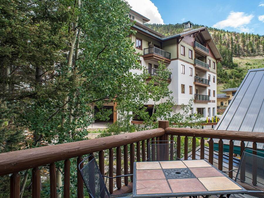 A wooden deck with a table and chairs, overlooking a multi-story building with balconies and a forested hillside in the background.