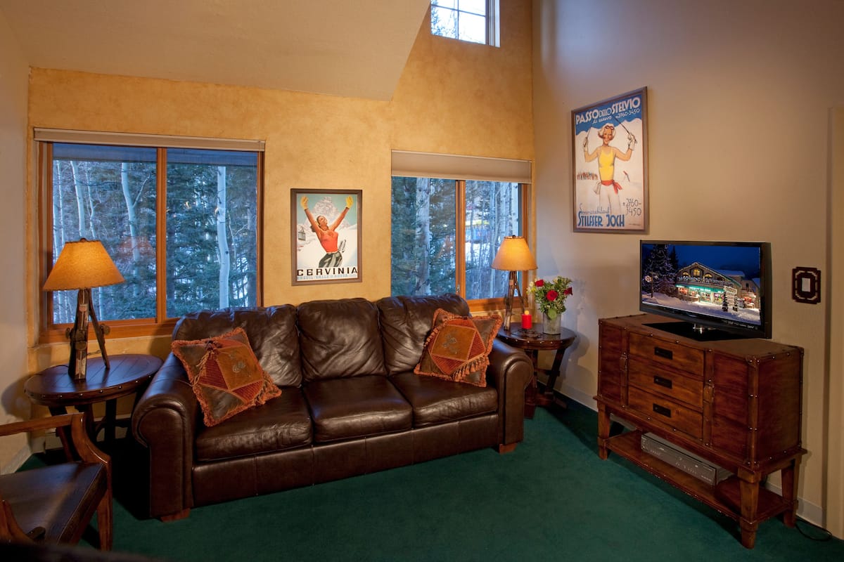 A living room with a brown leather sofa, two lamps, a television on a wooden stand, and framed posters on the wall.
