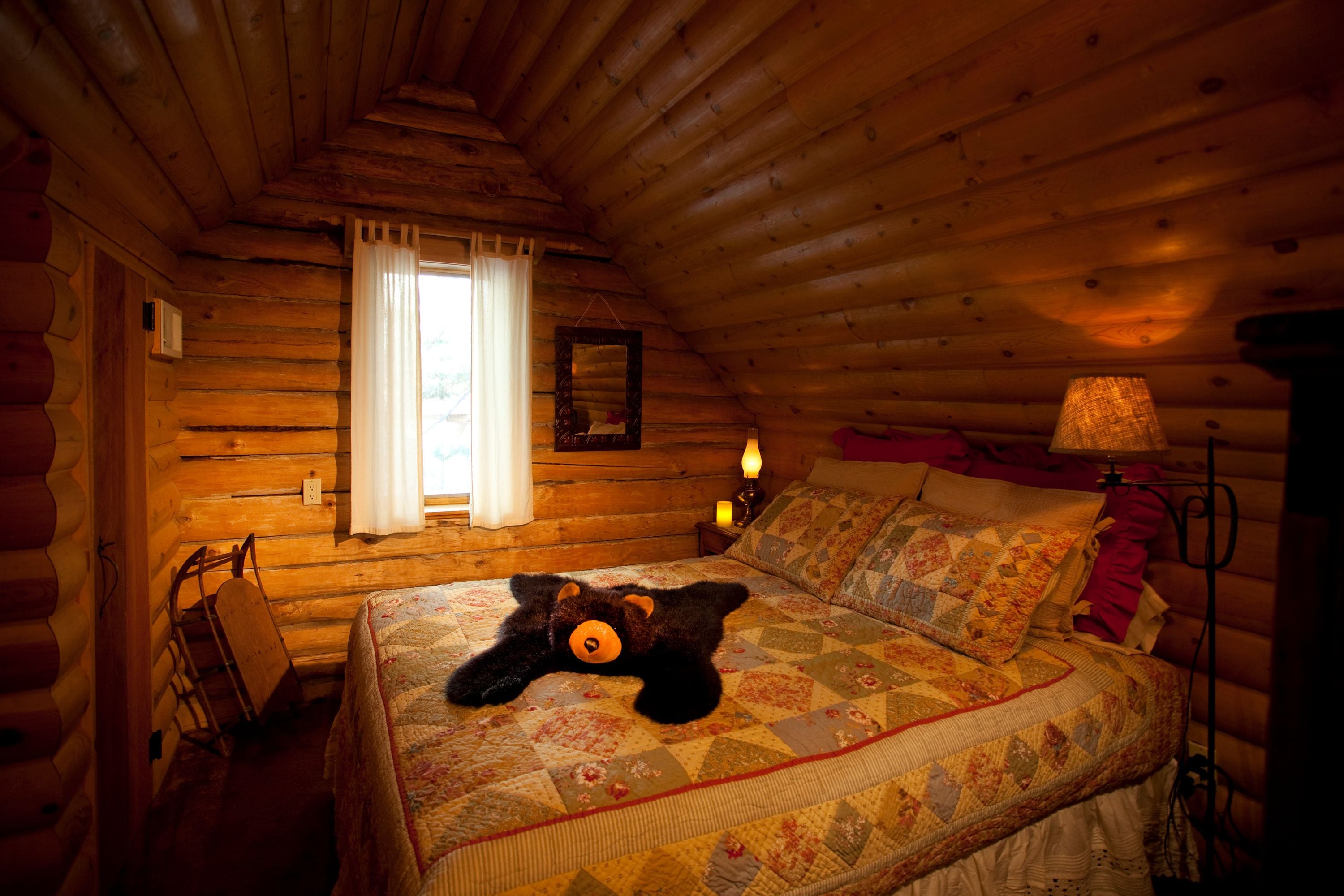 A cozy log cabin bedroom with a bed, a teddy bear, a lamp, and a window with curtains.