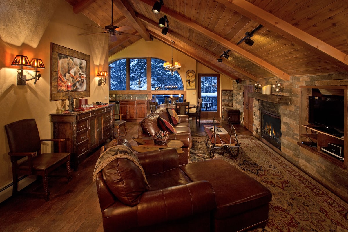 A spacious living room with a leather sofa, wooden ceiling beams, a fireplace, and a large window offering a view of trees.