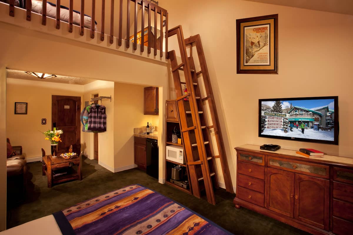 A room with a loft area, a television, a wooden ladder, a bed, a dresser, and a door leading to another room.