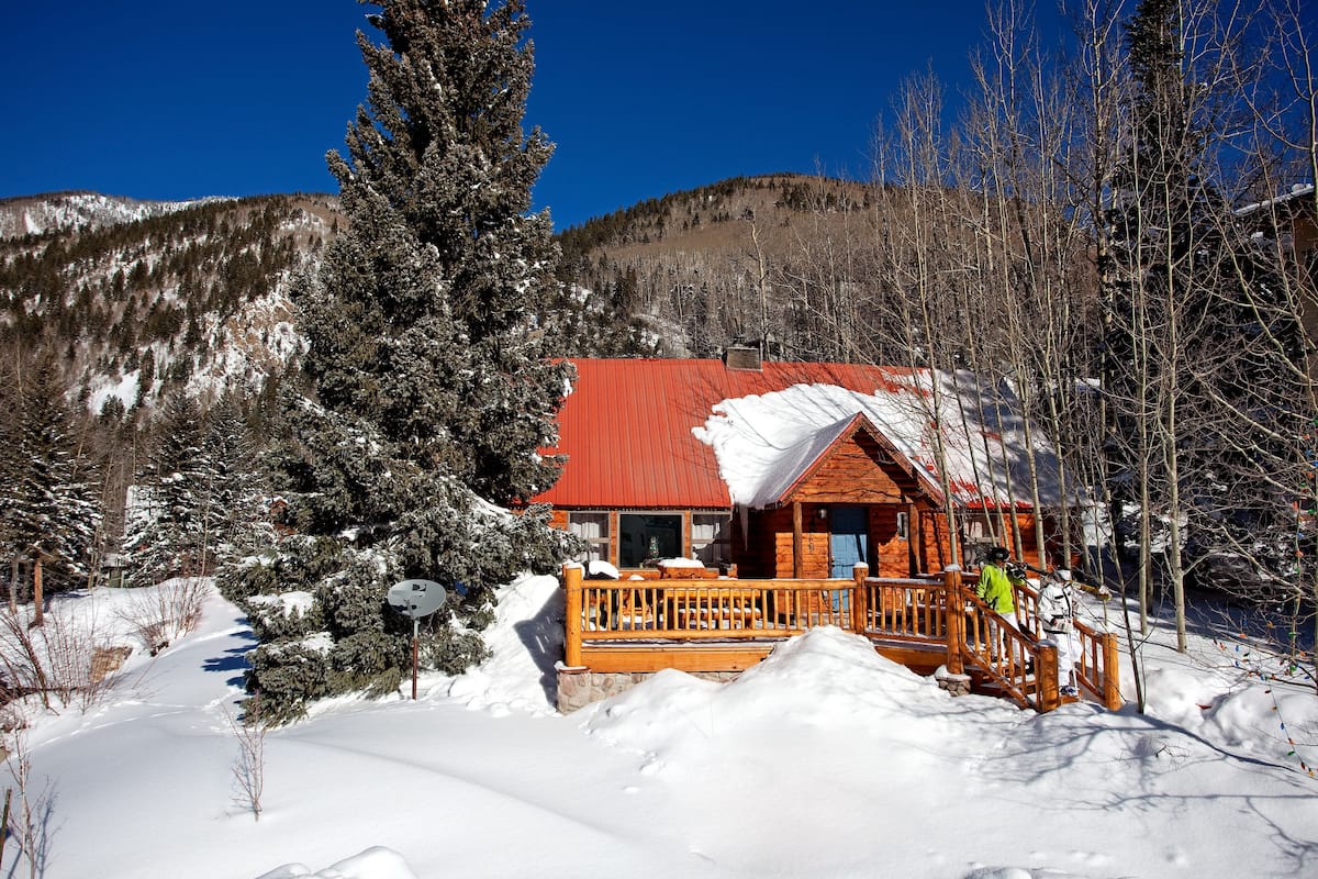 A wooden cabin with a red roof surrounded by snow-covered trees and mountains.