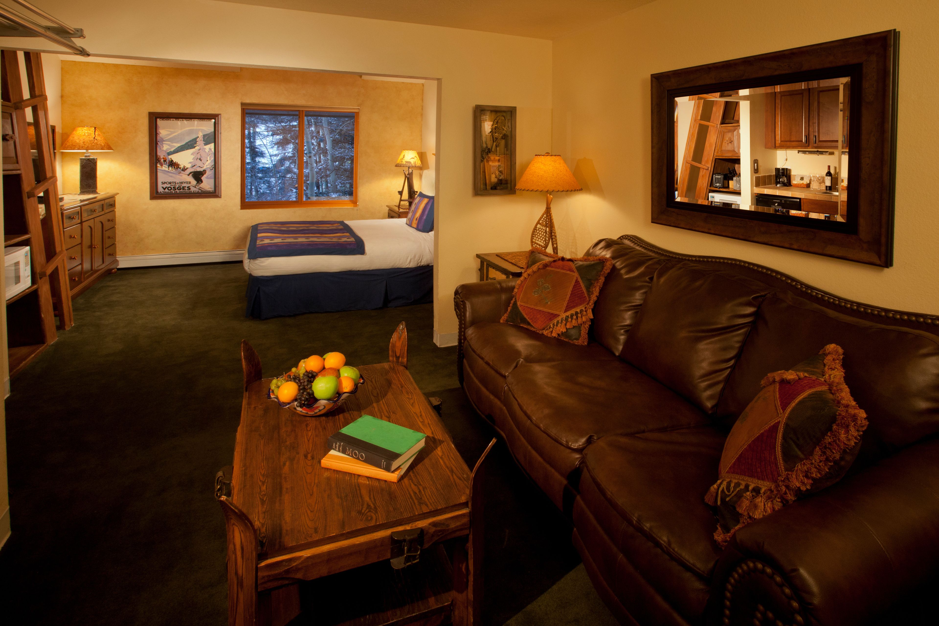 A hotel room with a bed, a wooden table with fruits and a book, a leather sofa, and a mirror reflecting the room.