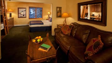 Habitación de hotel con cama, una mesa de madera con frutas y un libro, un sofá de cuero y un espejo que refleja la habitación.