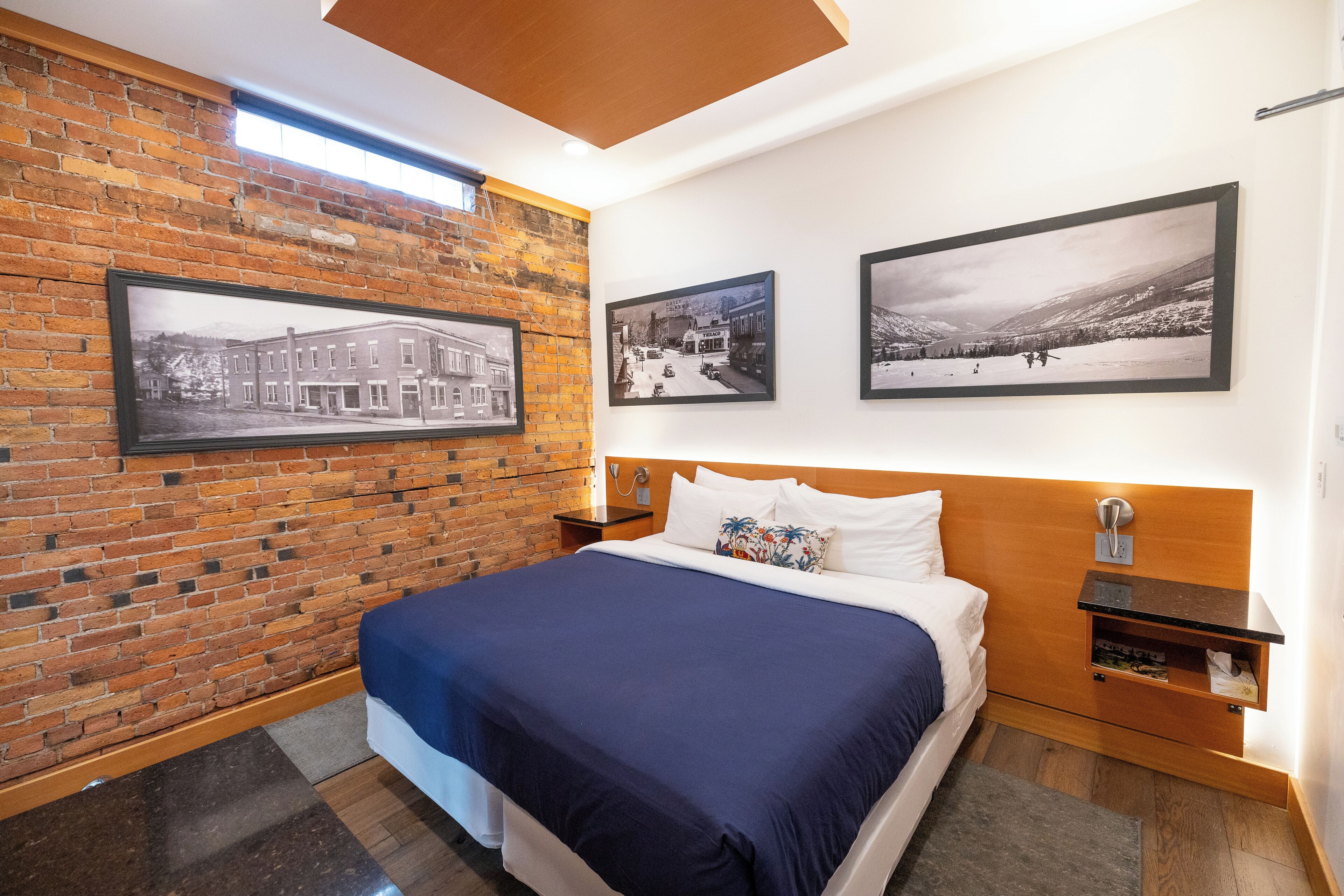 A modern hotel room with a brick wall, a large bed with a blue bedspread, and three framed photographs on the wall.