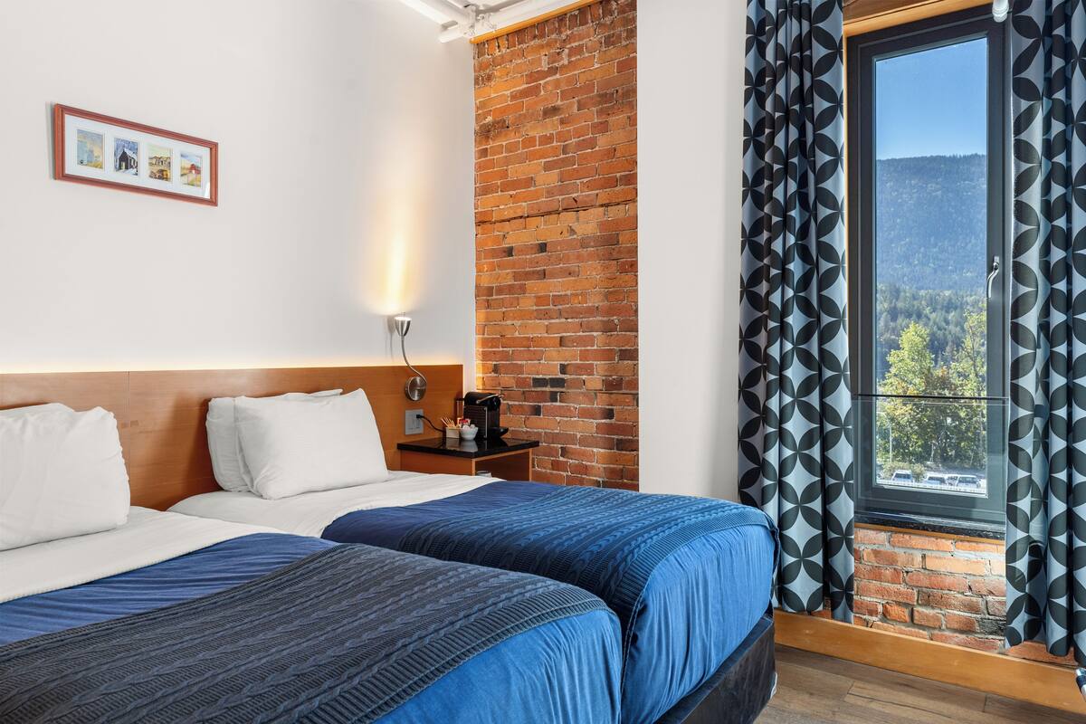 A hotel room with two beds, a brick wall, a window with curtains, and a framed picture on the wall.