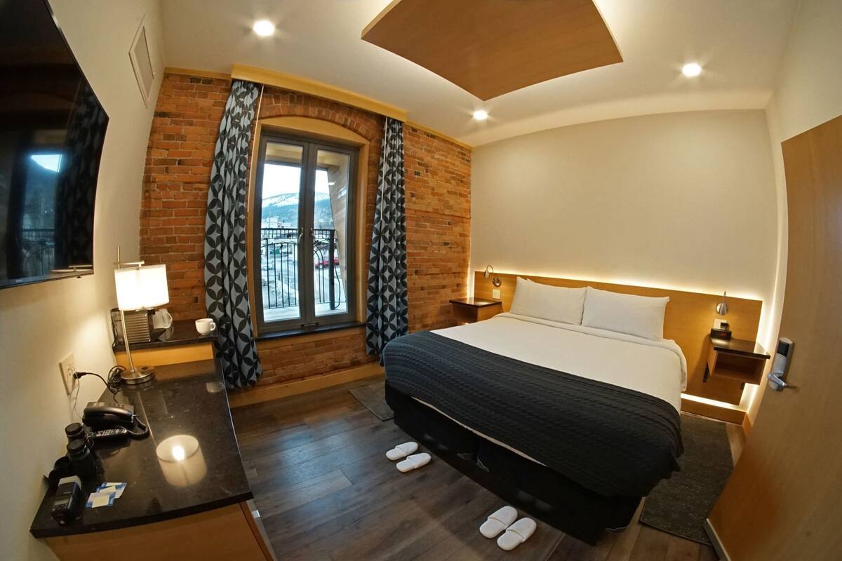 A hotel room with a bed, a desk with a lamp, a window with curtains, and a view of the outside.