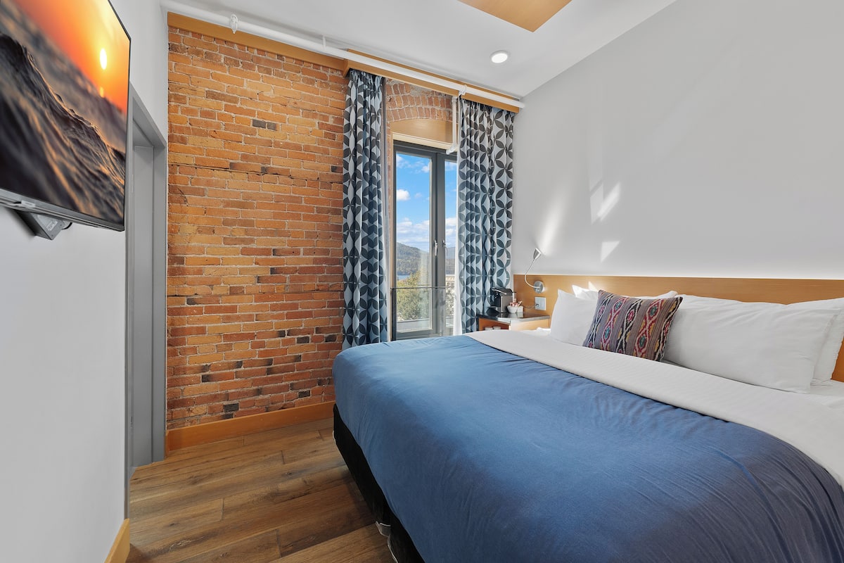 A hotel room with a brick wall, a large bed, a TV, and a window with curtains.