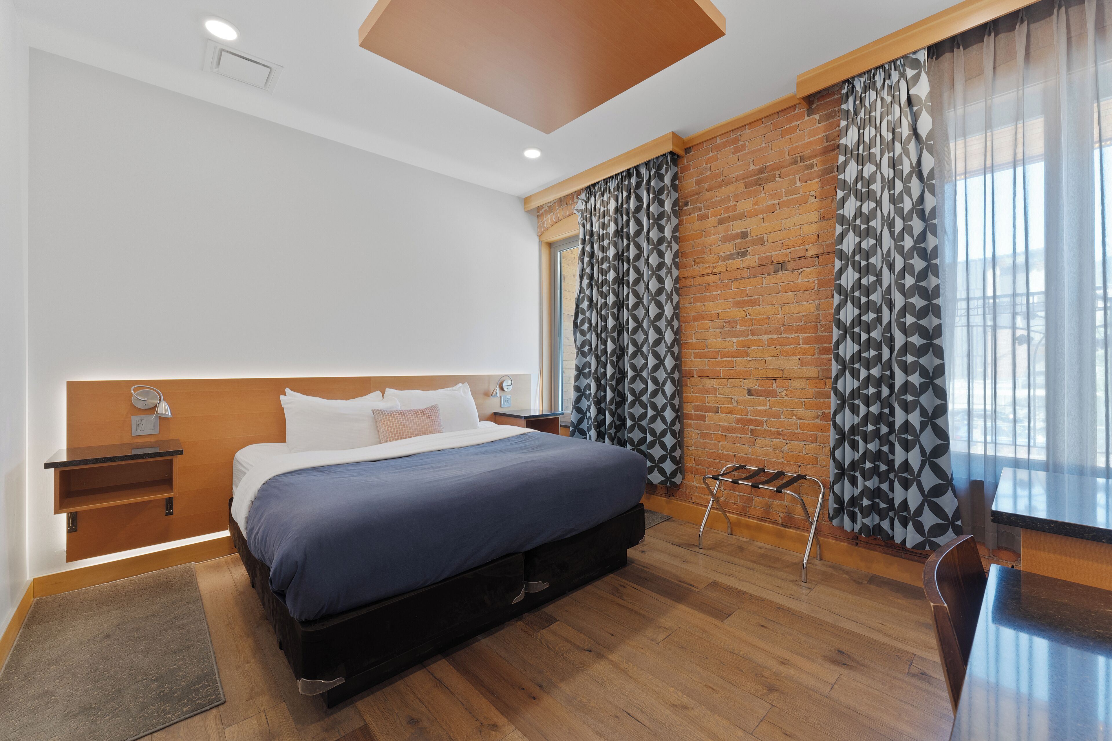 A modern hotel room with a large bed, wooden flooring, a brick accent wall, and a view of the city through sheer curtains.