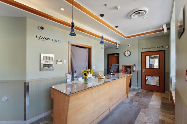 Un espace d’accueil avec un bureau en bois, une horloge et des panneaux indiquant «Savoy Hotel» et «Busaba Thai Cafe».
