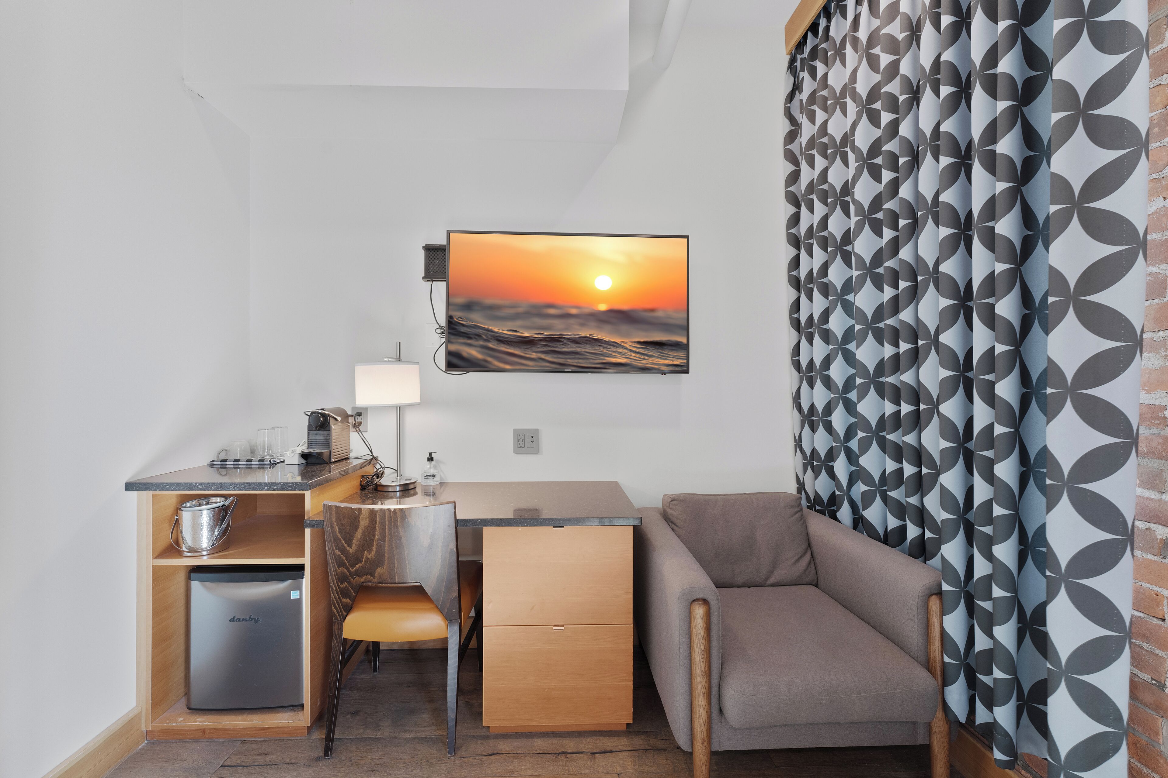 Une pièce compacte comprenant un bureau, une chaise et un canapé, ainsi qu’une petite kitchenette et un téléviseur fixé au mur, diffusant une image de coucher de soleil.