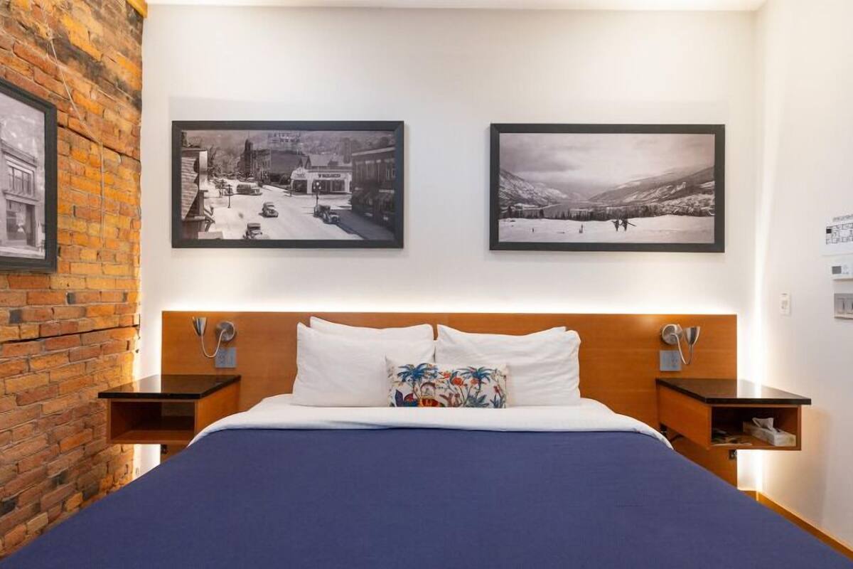 A hotel room with a brick wall, a bed with a blue bedspread, two framed pictures on the wall, and a wooden headboard.