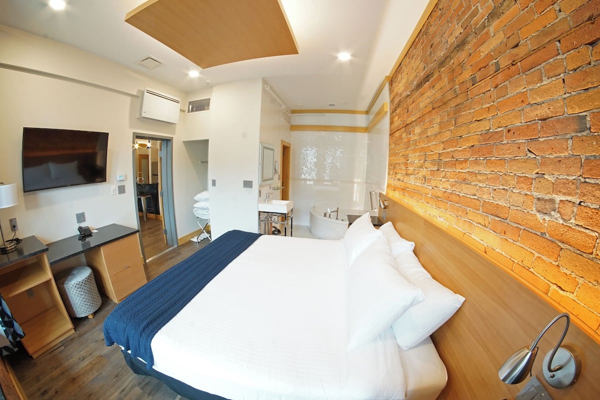 A hotel room with a large bed, a desk with a computer, a TV, and a brick wall.