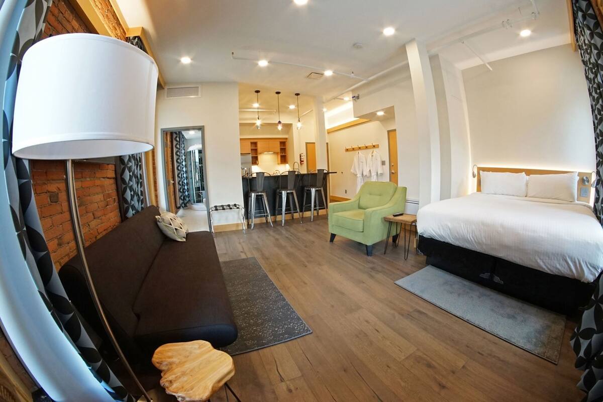 A modern hotel room with a bed, a sofa, a dining area, and a kitchen in the background.