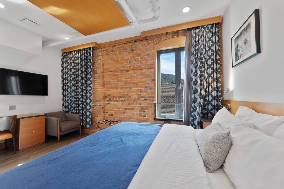 A modern hotel room with a large bed, a flat-screen TV, a wooden ceiling, and a brick wall.