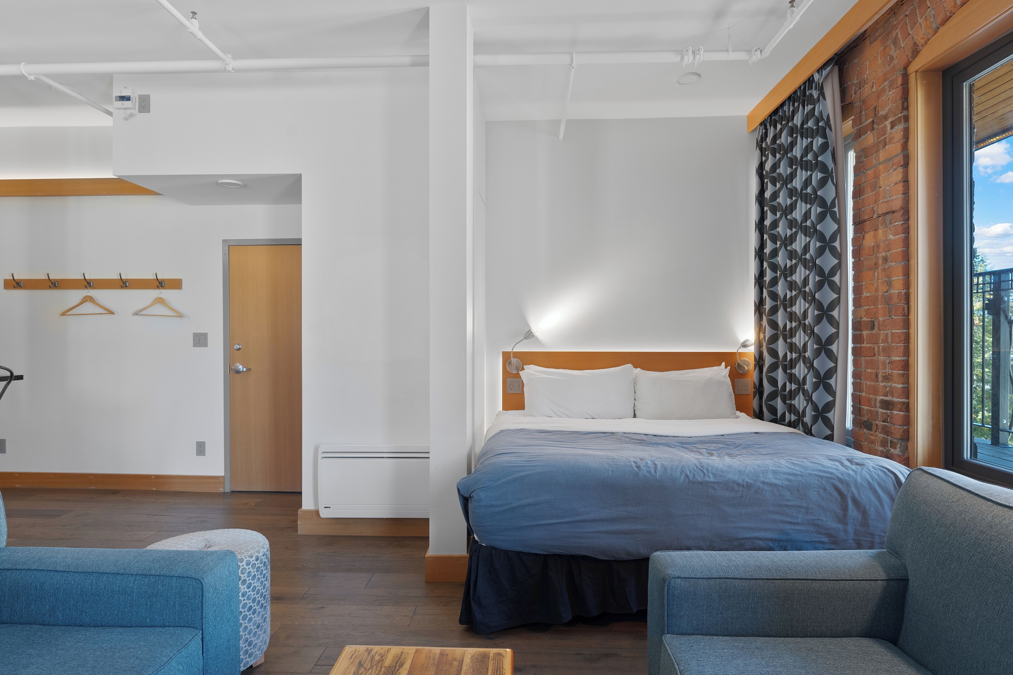 Ett modernt hotellrum med en stor säng, två fåtöljer, ett träbord och utsikt över omgivningarna genom ett fönster med mönstrade gardiner.