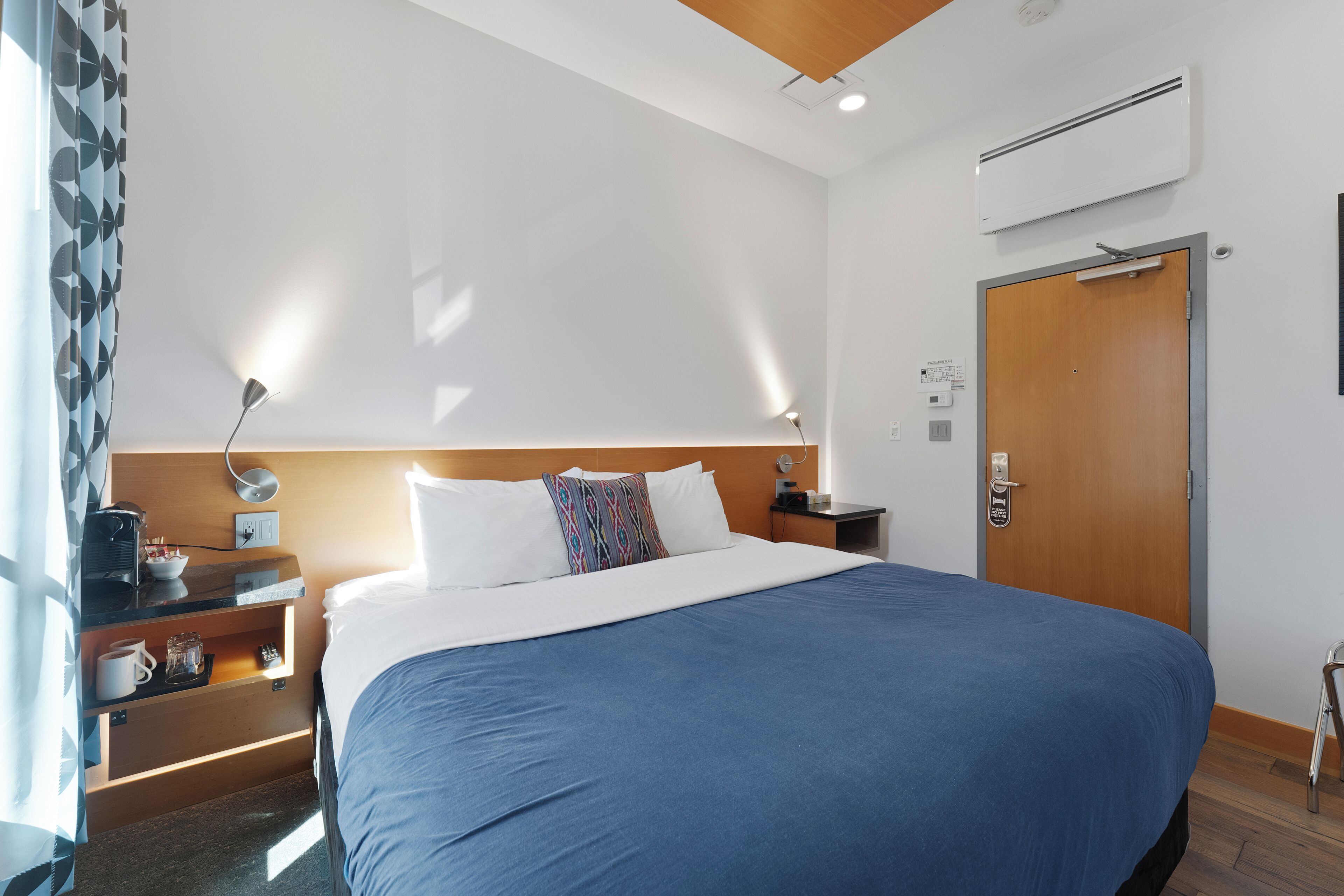 A hotel room with a large bed, a wooden headboard, a bedside table with a lamp, a nightstand, and a door with a handle.