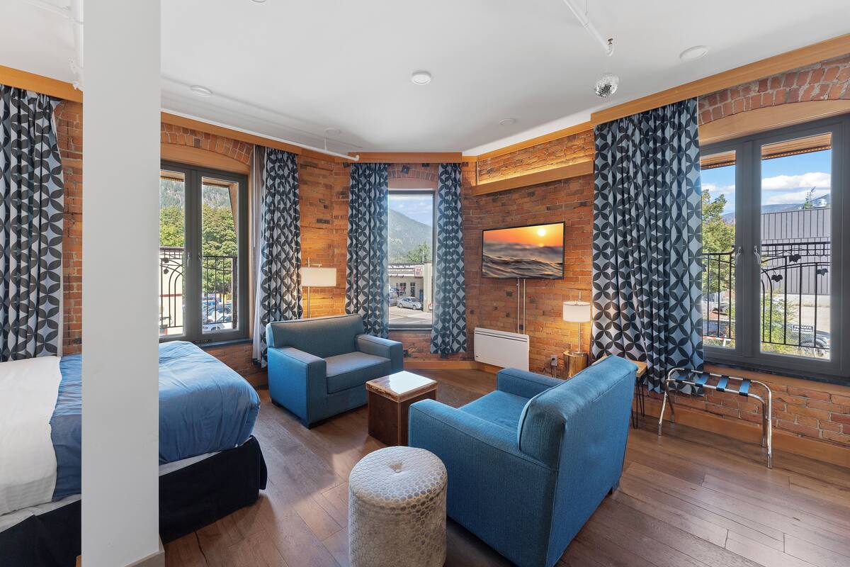 A modern hotel room with a bed, blue sofa, and armchair, large windows with a view of the outdoors, and a brick wall.