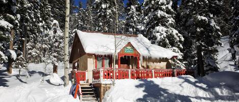 Una cabaña con barandilla roja y techo cubierto de nieve, rodeada de árboles cargados de nieve.