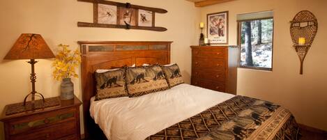 A bedroom with a wooden headboard, a bed with a patterned bedspread, a nightstand with a lamp, a dresser, and a window with a view of trees.
