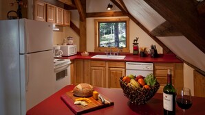 Uma cozinha com armários de madeira, bancada vermelha, janela com vista para árvores, geladeira, pia e tábua de corte com queijo e pão.