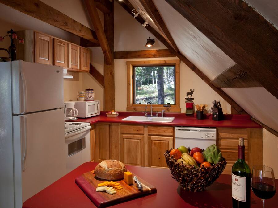 A kitchen with wooden cabinets, a red countertop, a window with a view of trees, a refrigerator, a sink, and a cutting board with cheese and bread.