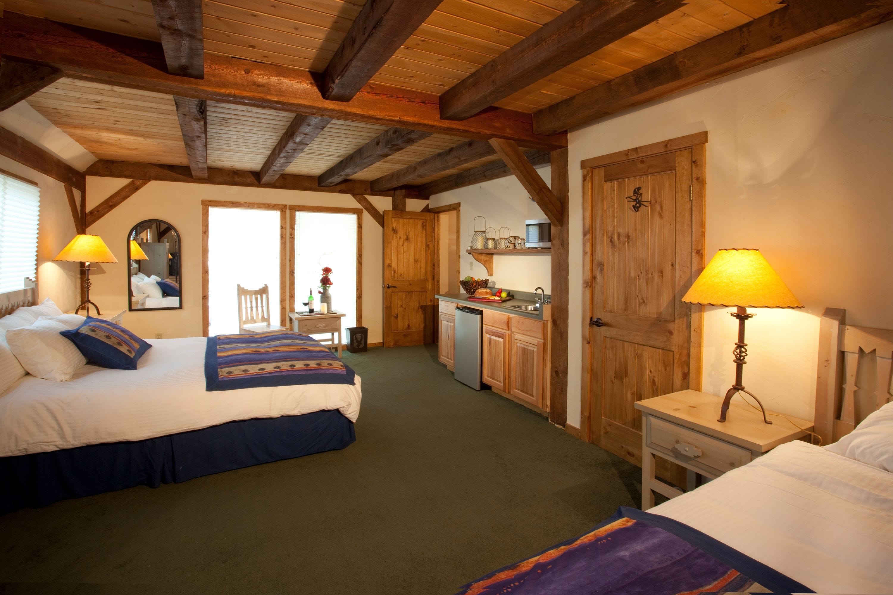 A bedroom with two beds, a wooden ceiling, a small kitchenette, and a mirror on the wall.