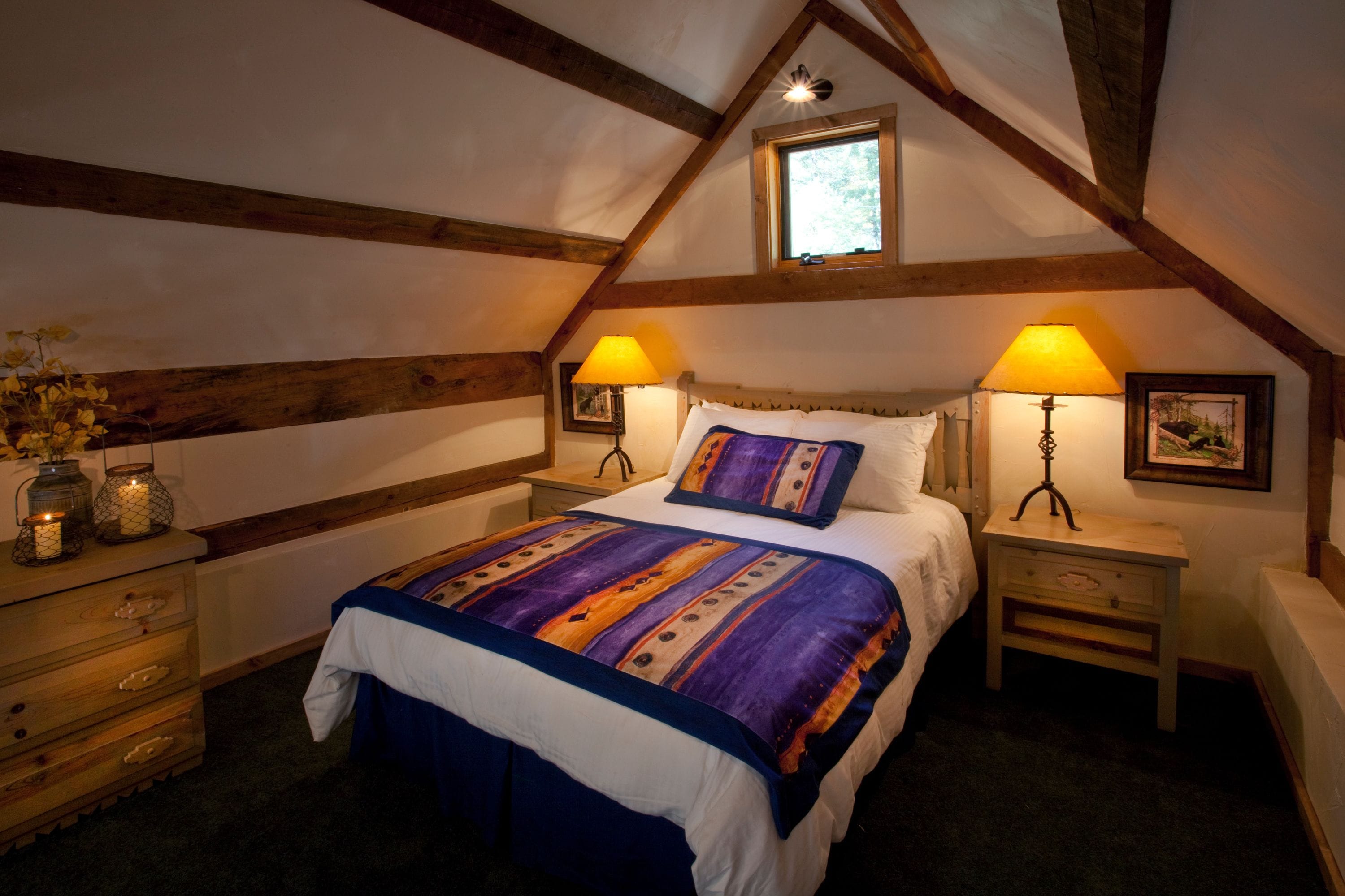 A bedroom with a sloped ceiling, wooden beams, a bed with a patterned bedspread, two bedside lamps, a nightstand with a lamp, a dresser, and a window with a view.
