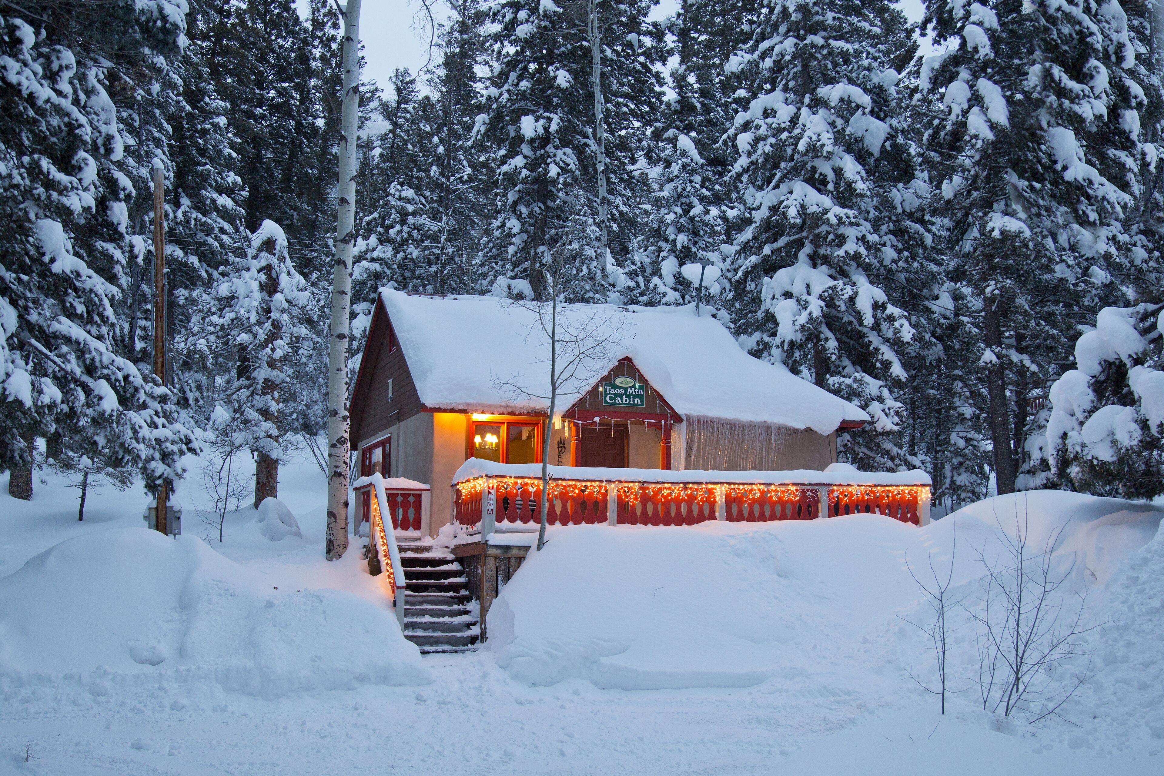 En stuga med ett snötäckt tak och en upplyst entré, omgiven av snötyngda träd.