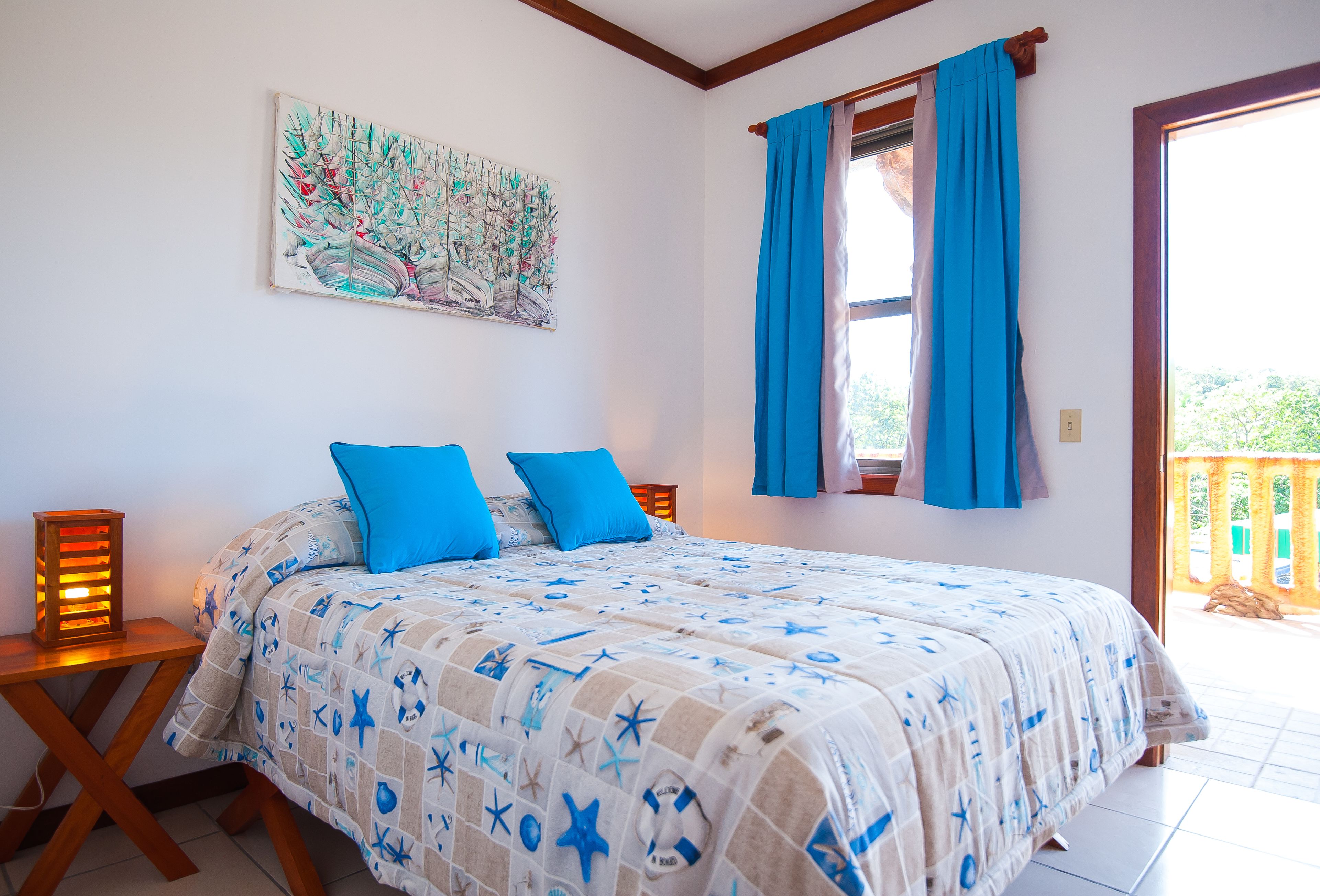 Un dormitorio con una cama, dos almohadas azules, una mesita de noche con una lámpara, un cuadro en la pared y una ventana con cortinas azules.