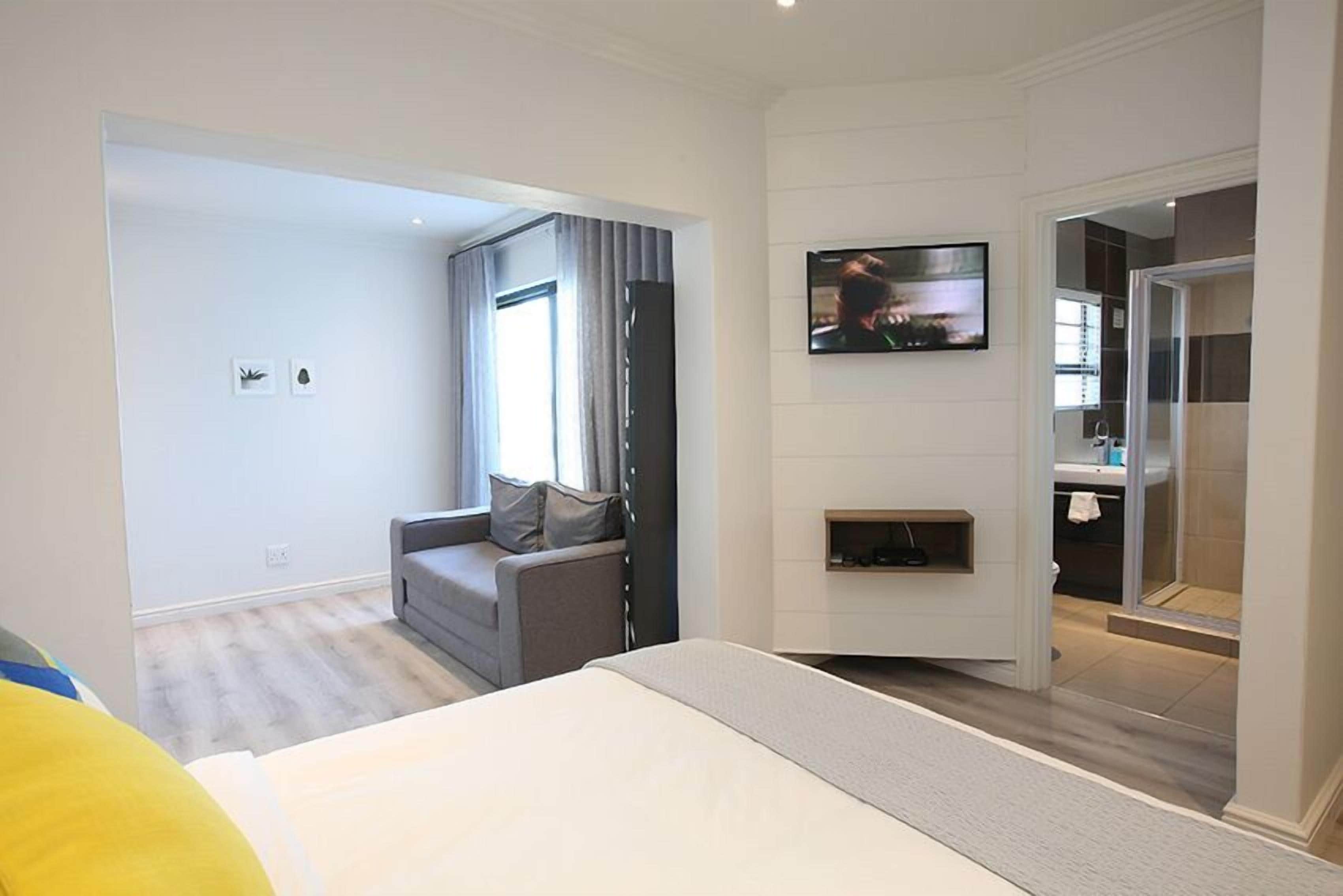 A modern hotel room with a large bed, a seating area with a sofa and armchairs, a flat-screen TV mounted on the wall, and a bathroom with a glass shower enclosure.