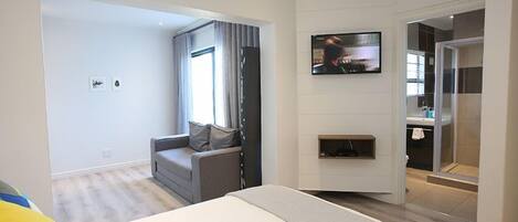 A modern hotel room with a large bed, a seating area with a sofa and armchairs, a flat-screen TV mounted on the wall, and a bathroom with a glass shower enclosure.
