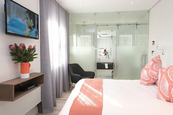 Habitación de hotel moderna con cama, televisor montado en la pared, escritorio con planta y zona de ducha visible a través de una puerta de vidrio.