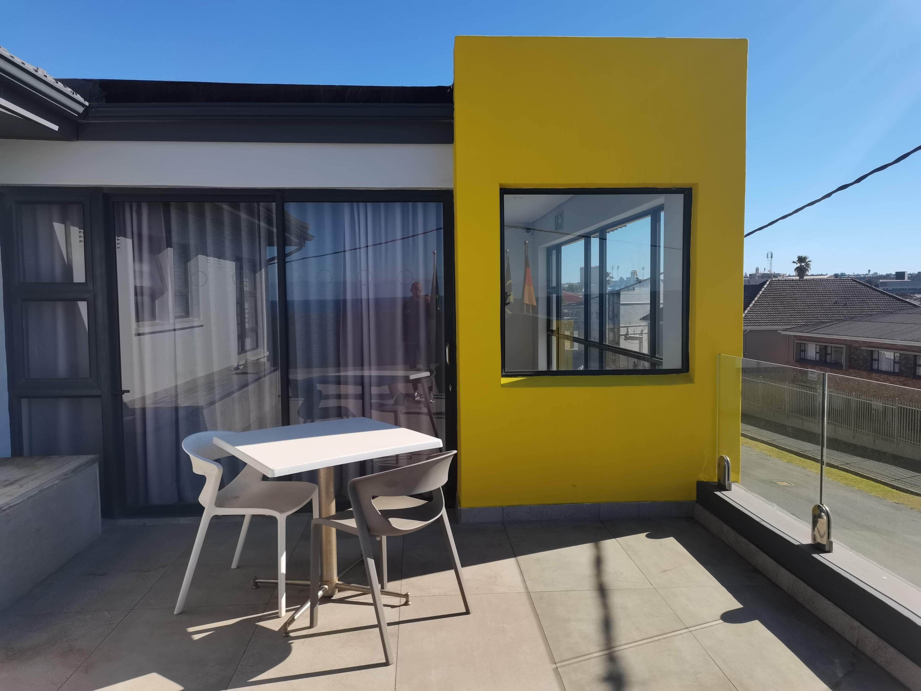 Un balcón con una mesa blanca y dos sillas, una pared amarilla y una ventana con vista a una zona residencial.