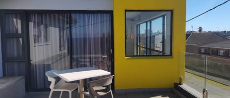 Un balcón con una mesa blanca y dos sillas, una pared amarilla y una ventana con vista a una zona residencial.