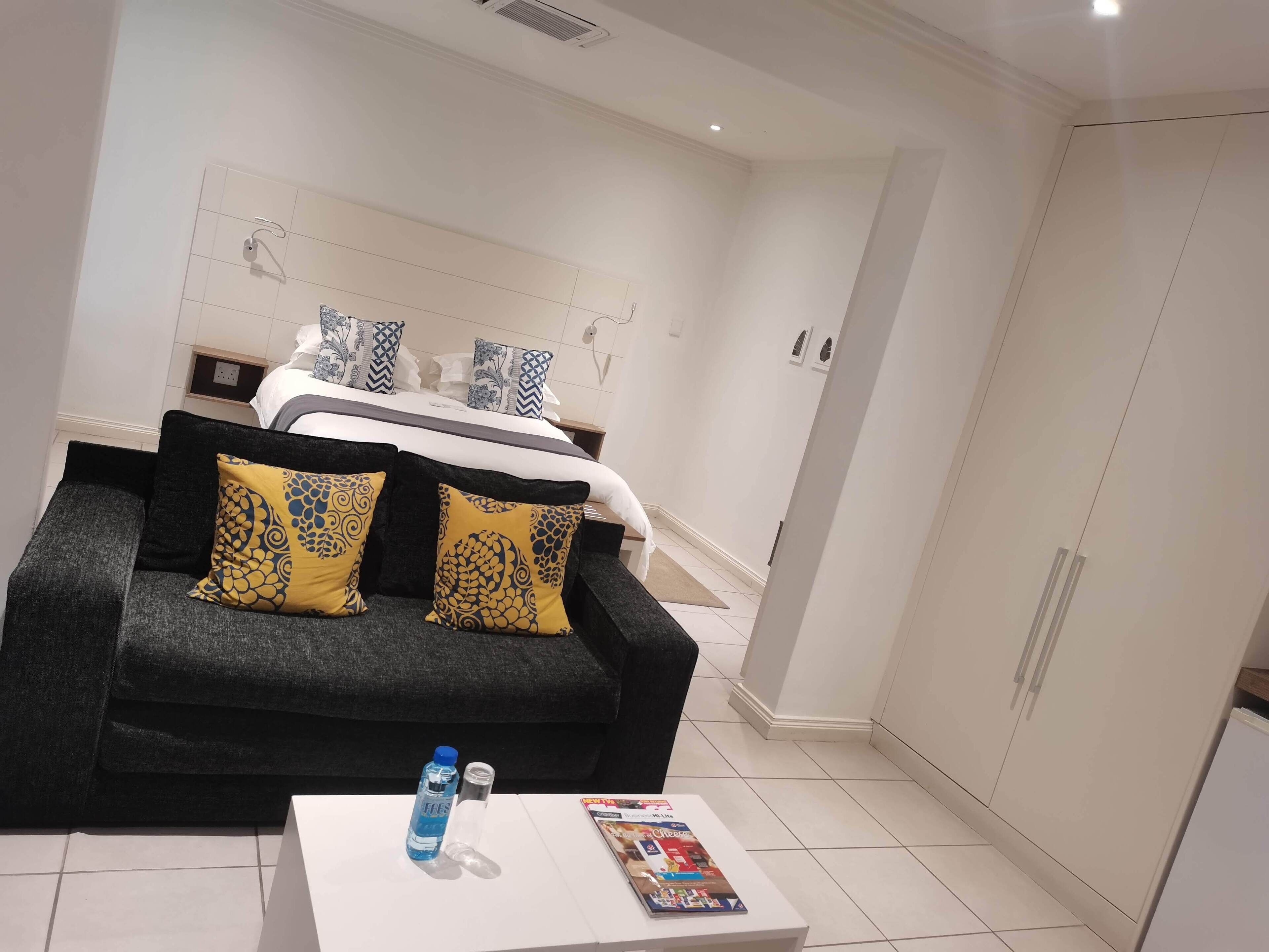 A hotel room with a bed, a black sofa with yellow and black patterned cushions, a white coffee table with a magazine and a water bottle, and a wardrobe with a mirror.