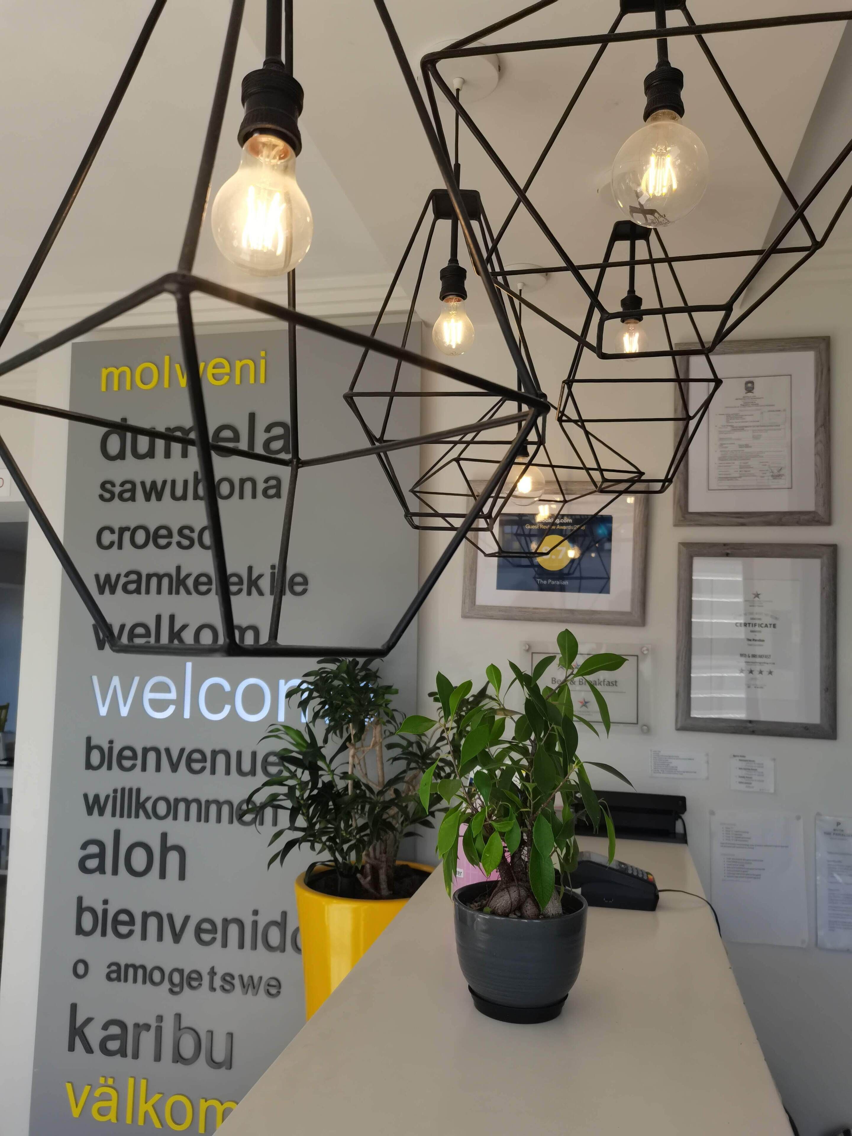 A reception area with a modern geometric light fixture, a welcome sign with multiple languages, and potted plants.