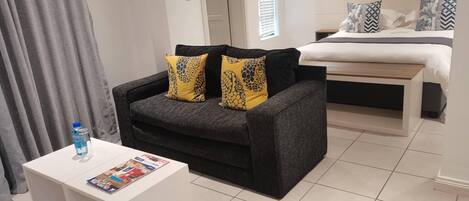 Una habitación moderna con un sofá gris, dos cojines amarillos estampados, una mesa de centro blanca con revistas, una cama con almohadas estampadas y una ventana con persianas.