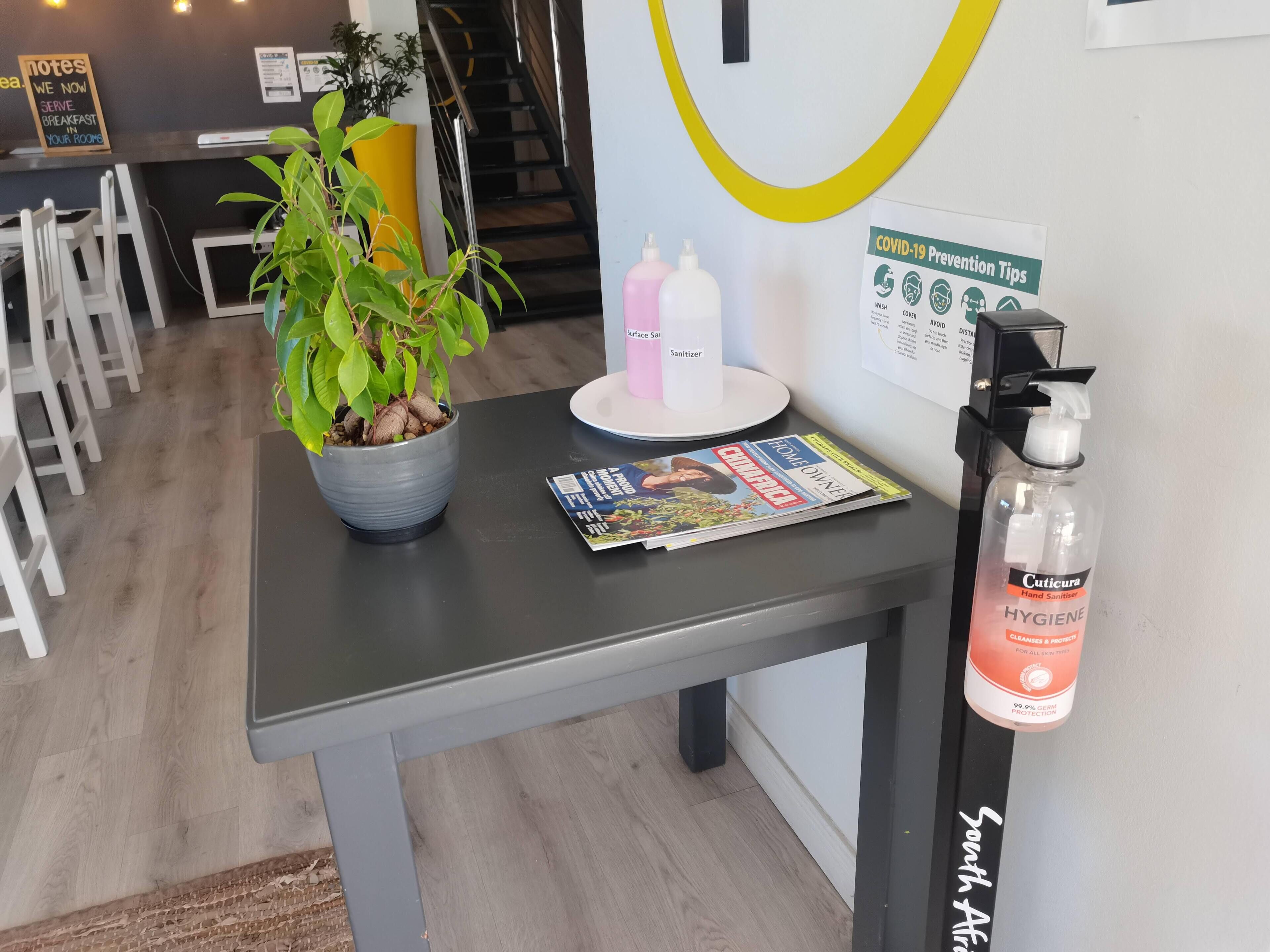 A reception desk with a magazine, a potted plant, a hand sanitizer dispenser, and a COVID-19 prevention sign.