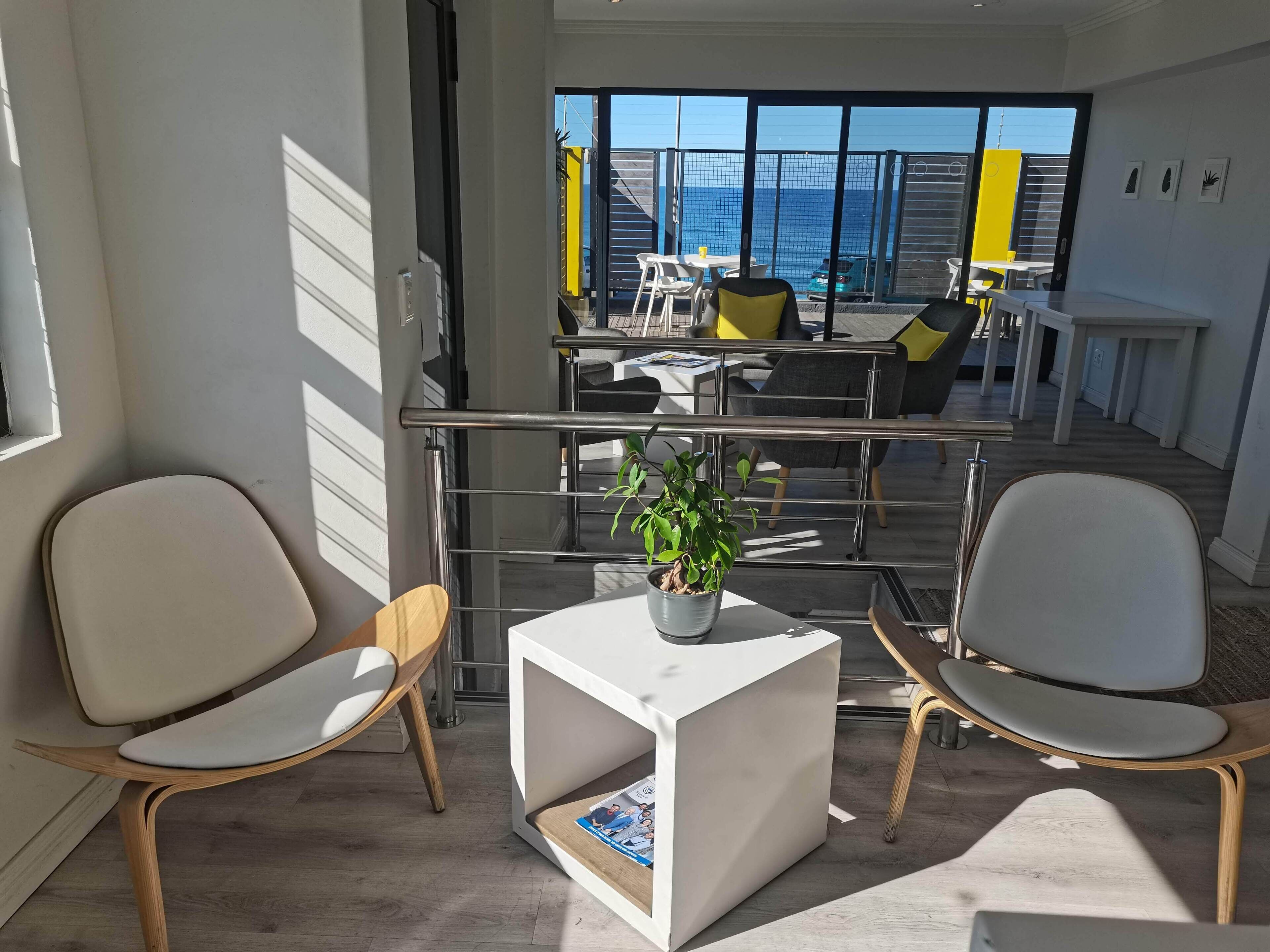 A modern, well-lit room with two white chairs, a small white table with a plant, and a view of the ocean through large windows.
