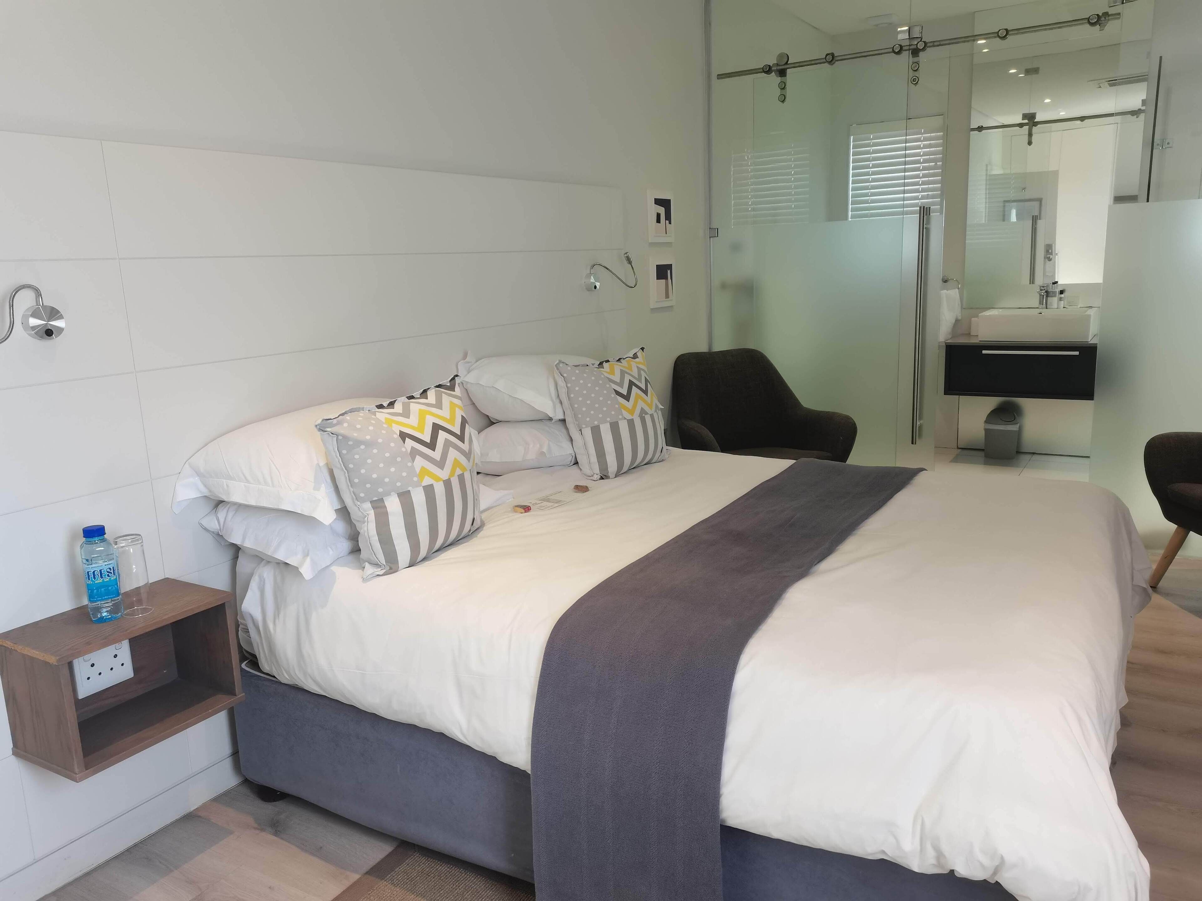 A neatly made bed with pillows, a bedside table with a bottle and glass, a chair, and a bathroom with a mirror and sink.