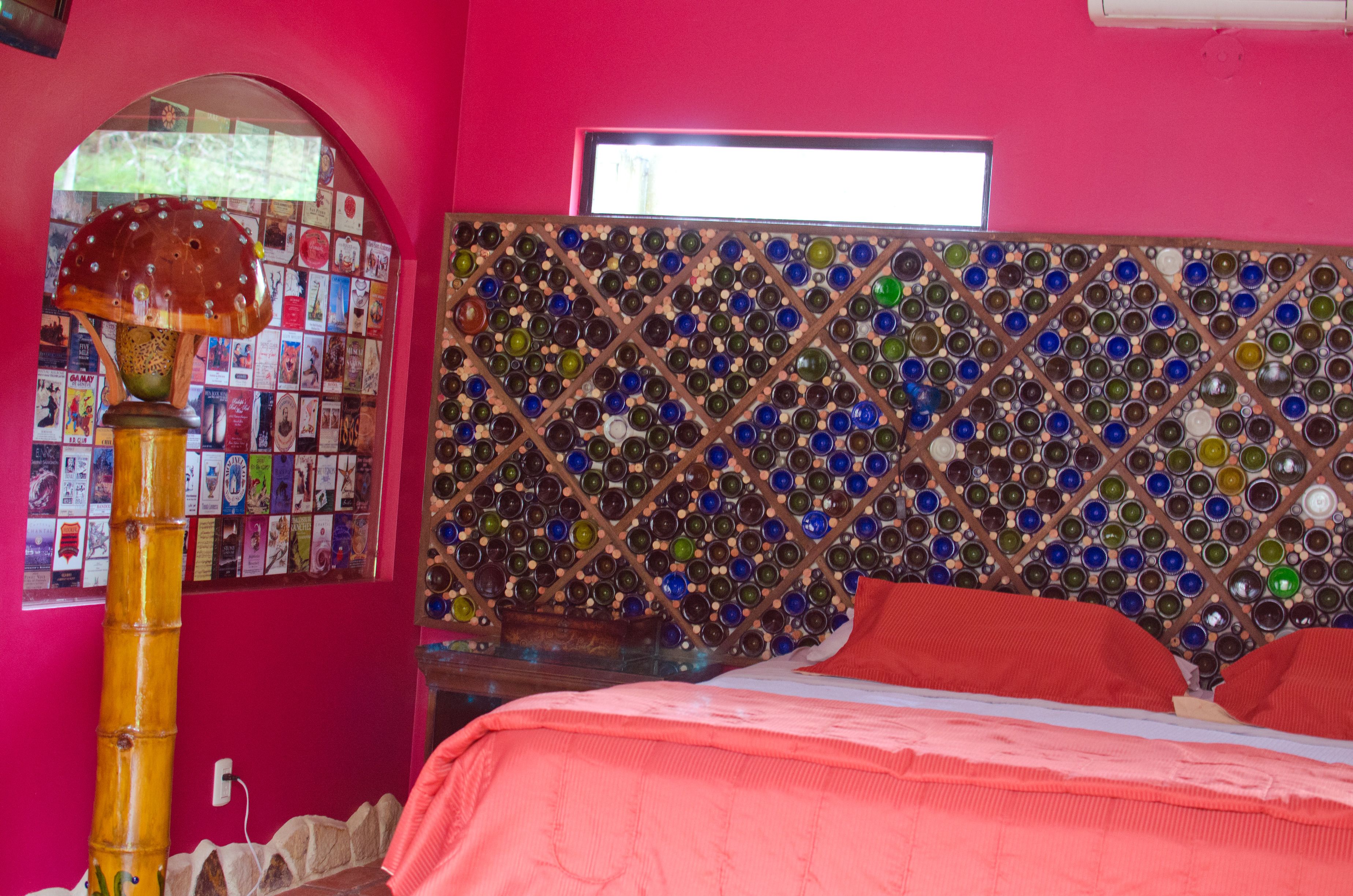 A room with a wall mosaic of glass bottles, a bed with red bedding, and a bamboo lamp.
